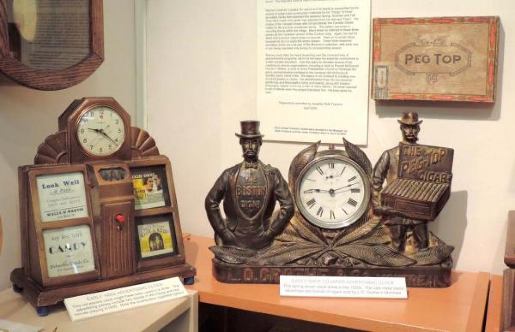 Two 1920s/30s Canadian advertising clocks: electric "diner" clock with 1930 movie ads and red push-button cigarette lighter;  cigars promotion windup clock in cast metal stand.  Note the matching old cigar box on the wall.